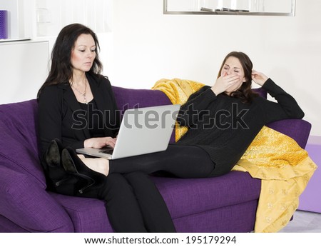 woman sitting on couch with a laptop while another woman is bored and yawns