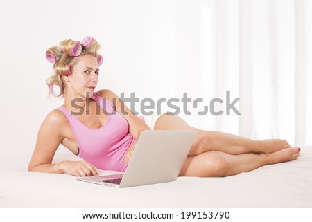 a blond woman with  pink curlers laying in bed with a laptop
