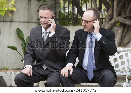two businessmen sitting outside on a bench and talking on the phone