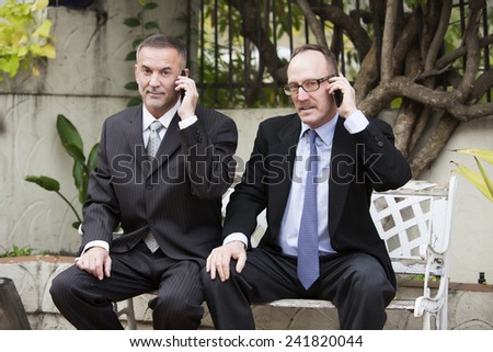 two businessmen sitting outside on a bench and talking on the phone