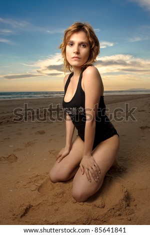 Sexy woman with black tank top on the beach on her knees at sunset with amazing sky on the background