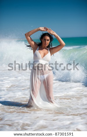 Brunette woman with a white transparent and wet dress at the beach on the water posing.