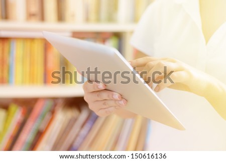 Female hands, holding tablet computer, bookshelves at background - library or college class or hospital interior, bright warm sunlight effect.