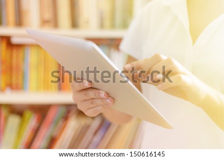 Female hands, holding tablet computer, bookshelves at background - library or college class or hospital interior, bright warm sunlight effect.
