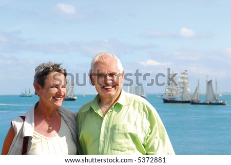 senior retired smiling couple walking outdoors with scenic ocean and sailboats background