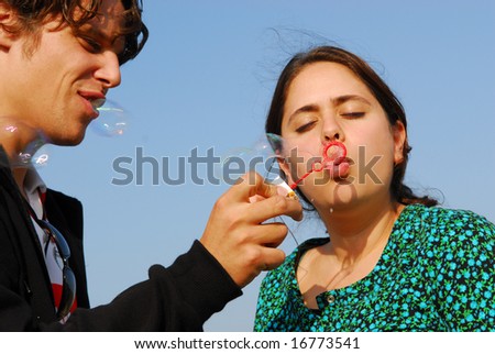 Concept: stay young. Man in his twenties lets his girlfriend blow soap bubbles on blue sky background