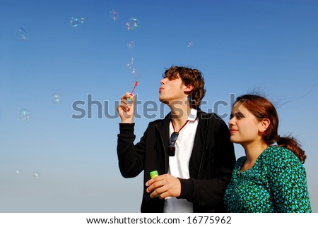 Concept: stay young! Man in his twenties blowing soap bubbles for his girlfriend on blue sky background
