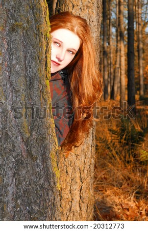 Caucasian girl with long red hair, peeking from behind a tree in an autumn colored forest