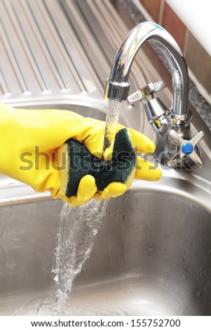 Hand In A Rubber Glove Holding A Cleaning Sponge Under A Running Tap In A Stainless Steel Sink