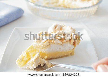 a slice of lemon meringue pie on a plate, with the pie dish in the background, and a piece of pie on a fork ready to be eaten.