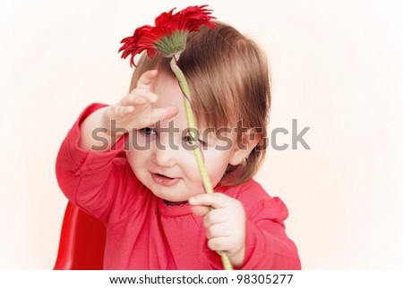 Smiling toddler on a pale background holding a red flower above her head.