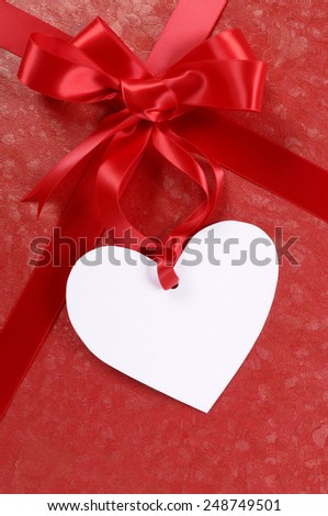 White heart shape valentine gift tag on a red gift ribbon and bow background.  Space for copy.