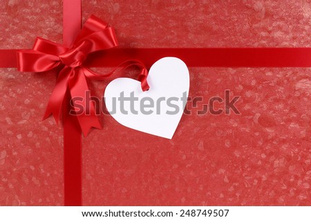 White heart shape valentine gift tag on a red gift ribbon and bow background.  Space for copy.