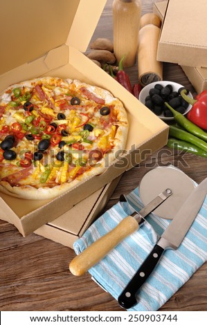 Freshly baked Pizza in a delivery box surrounded by various ingredients on a wood table or worktop.