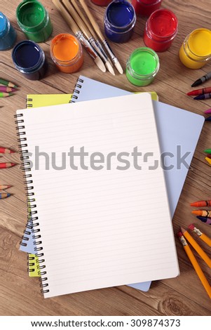 Blank writing book on a school desk with various paints, pencils and crayons, vertical format