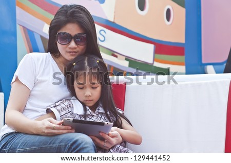 Little asian girl and mom enjoy tablet PC in shopping mall.