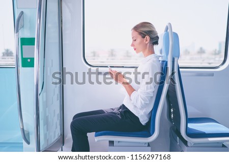 Enjoying travel. Young pretty woman traveling by the metro train sitting near the window using smartphone.