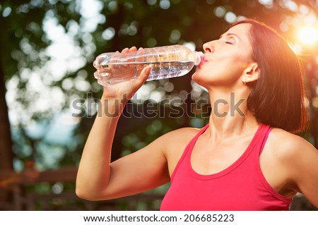 Senior athletic woman drinks water from a bottle after running in the park.