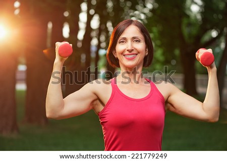 Portrait of cheerful aged woman in fitness wear exercising with red dumbbells in park.