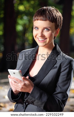 Staying connected. Beautiful young woman in formalwear holding the mobile phone, looking at camera and smiling while standing outdoors