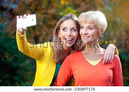Portrait of smiling caucasian middle aged mother and her blonde adult daughter are doing selfie by white mobile phone outdoors