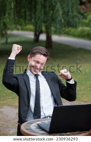 Business winner. Happy young man in suit and tie keeping arms raised and expressing positivity while sitting at the  outdoors cafe working with laptop with park in the background