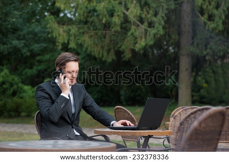 Busy working. Confident young caucasian businessman in formalwear working on laptop and talking on the mobile phone while sitting at the sidewalk cafe.