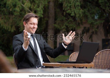 Business winner. Happy young caucasian man in suit and tie keeping arms raised and expressing positivity while sitting at the  outdoors cafe working with laptop with park at the background