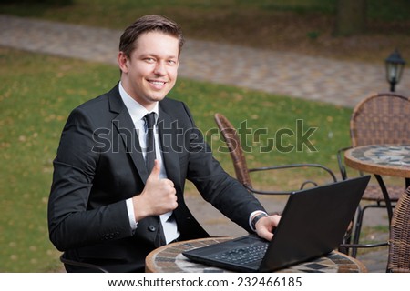 Good job! Happy young businessman in suit and tie showing his thumb up and smiling while working on laptop at cafe outdoors