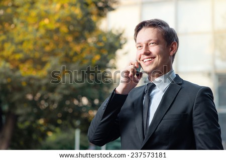 Confident business talk. Cheerful smiling caucasian young man in formal wear talking on the mobile phone and looking away while standing outdoors and against building structure outdoors