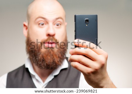 Shocking news. Surprised bearded man holding mobile phone and looking at it while standing against grey background. Focus on the phone.