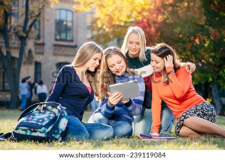 Surfing the net together. Four happy young caucasian woman looking at tablet pc and smiling while  they rest on grass at campus together