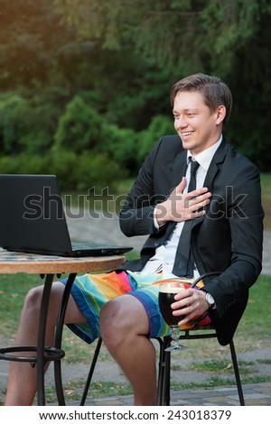 Work and relax. Stay connected and drink a beer! Caucasian businessman dressed in suit and shorts working with laptop, talking by skype at the park cafe outdoors