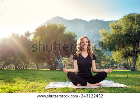 Happy morning yoga. Cheerful Young woman in lotus pose in the park with mountains view. Practicing yoga with trees and sun ray in the background