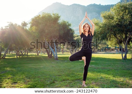 Happy young woman standing in yoga pose on the grass in the park. Practicing yoga with trees, mountains and sun ray in the background