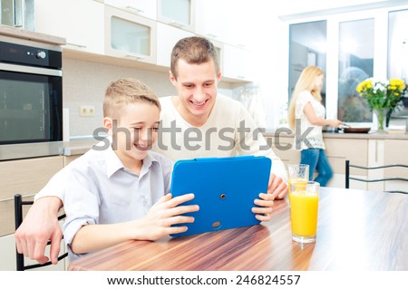 Happy family and digital technology. Smiling boy and his father using a tablet pc in a kitchen while mother cooking dinner at the background.