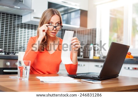 stressed over bills. Surprised young woman looking at her financial debts in the kitchen at home wearing orange pullover sitting at dining table.