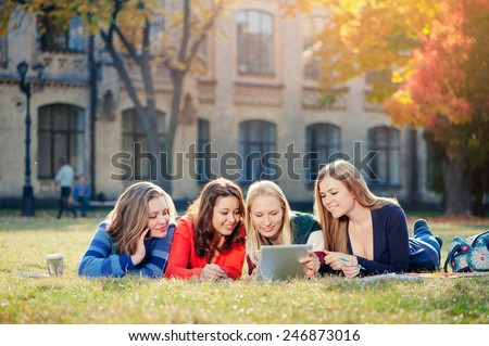 Surfing the net together. Four happy caucasian young woman looking at tablet pc and smiling while  they rest on grass at campus together