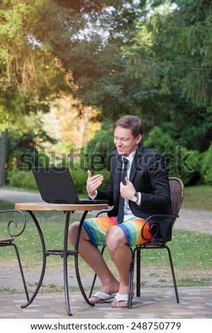 Work and relax. Stay connected! Caucasian businessman dressed in suit and shorts working with laptop, talking by skype at the park cafe outdoors