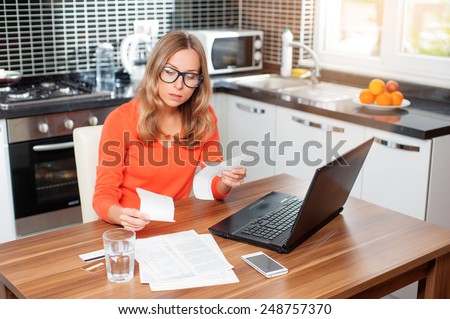 stressed over bills. portrait of a young  woman using a laptop computer sitting at her kitchen holding utility bills and bank statements being thoughtful and worried. Home kitchen interior