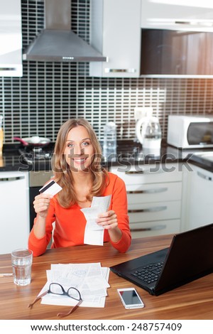 Attractive young smiling Woman holding credit card paying bills online from home while sitting in he kitchen