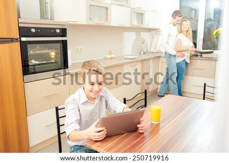 Smiling boy using digital tablet in the kitchen while his parents cooking dinner at the background.
