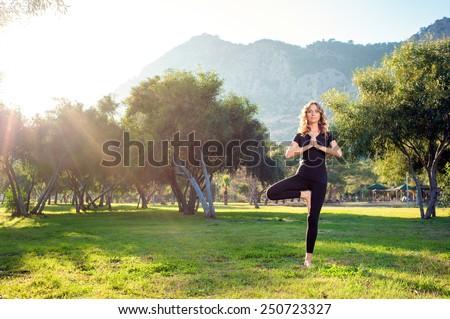 Practicing yoga in the morning, with trees, mountains and sun ray in the background. Attractive young caucasian woman standing in yoga pose on the grass.