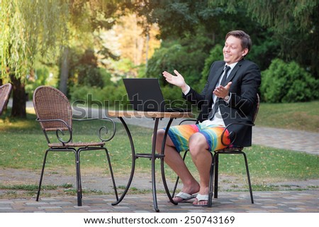 Work and relax. Stay connected! Businessman dressed in suit and shorts working with laptop, talking by skype at the park cafe outdoors