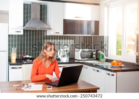 Business woman in orange pullover working from her dining table. Home kitchen in the background.