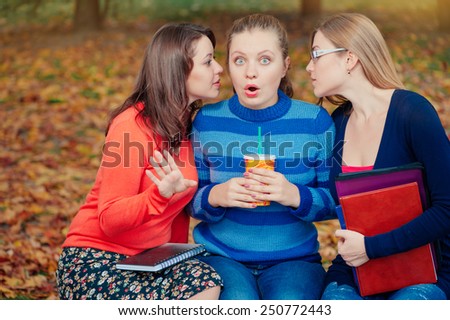 Shocking Secrets. gossip, young women whispering to the friend\'s ear while sitting on bench in autumn park.