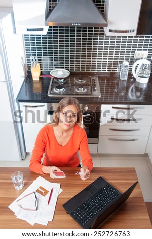 Happy young caucasian woman with laptop using credit card and phone in kitchen at home. top view