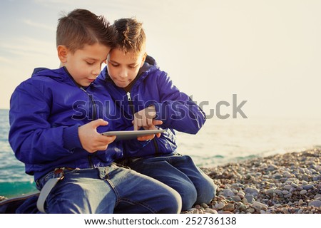 Two happy caucasian kids, brothers, playing together with tablet pc sitting outdoors at pebble beach against the sea