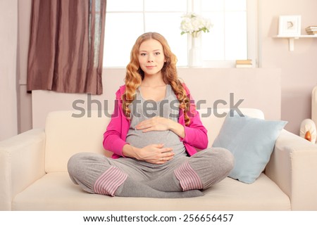 Keeping calm for her unborn baby. Beautiful pregnant woman sitting in lotus position on couch and smiling