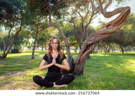 Morning meditation. Young caucasian woman with closed eyes in lotus pose in the park sitting on green grass under the tree.
